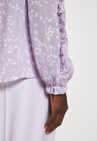 Blouse lavande avec taches blanches abstraites et poignet de manche à volants portée avec un pantalon lavande clair sur une personne à la peau foncée.