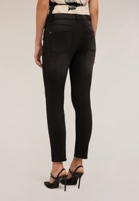 Mujer vestida con jeans ajustados negros y zapatos de tacón alto tipo slingback negros, de pie frente a un fondo neutro.