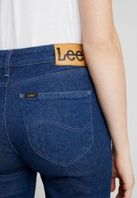 Jeans de denim azul oscuro con una etiqueta de cuero marrón que presenta "Lee" en la cintura, con costuras visibles y detalles de los bolsillos.