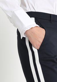 Granatowe spodnie z białymi paskami po bokach, wykonane z gładkiego materiału, w połączeniu z białą bluzką z falbaniastymi mankietami, z ręką w kieszeni.