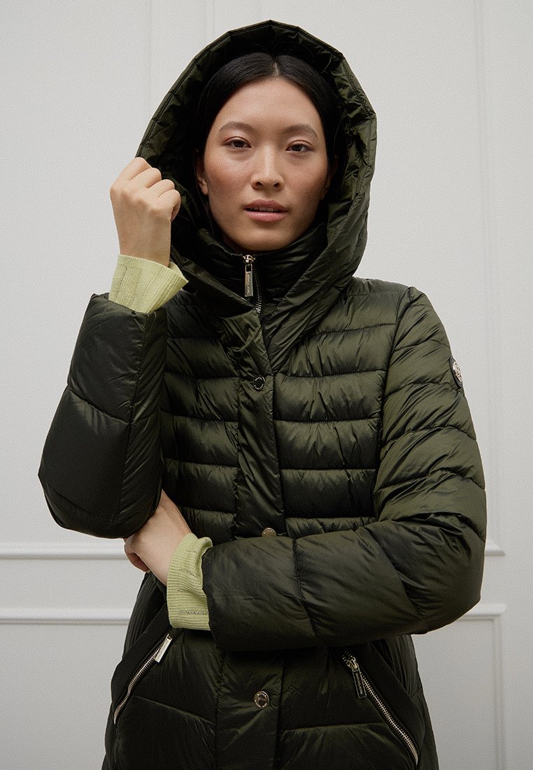 Frau trägt eine dunkelgrüne Steppjacke mit Kapuze, hält die Kapuze mit einer Hand, vor neutralem Innenhintergrund.