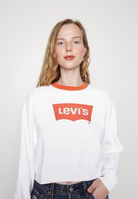 Fehér, rövidített pulóver narancssárga nyakkal és egy nagy vörös Levi's logóval. Az anyag lágy és texturált, alkalmas hétköznapi viseletre.