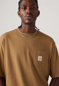 Tricou din bumbac maro cu guler rotund, mâneci scurte și un buzunar pe partea stângă, dotat cu o etichetă albă cu text. Textură netedă.