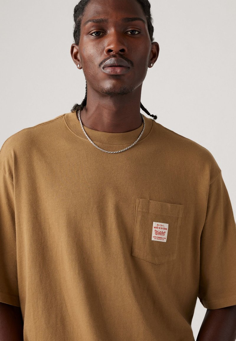 T-shirt in cotone marrone con collo a giro, maniche corte e taschino sul lato sinistro con un'etichetta bianca con testo. Texture liscia.