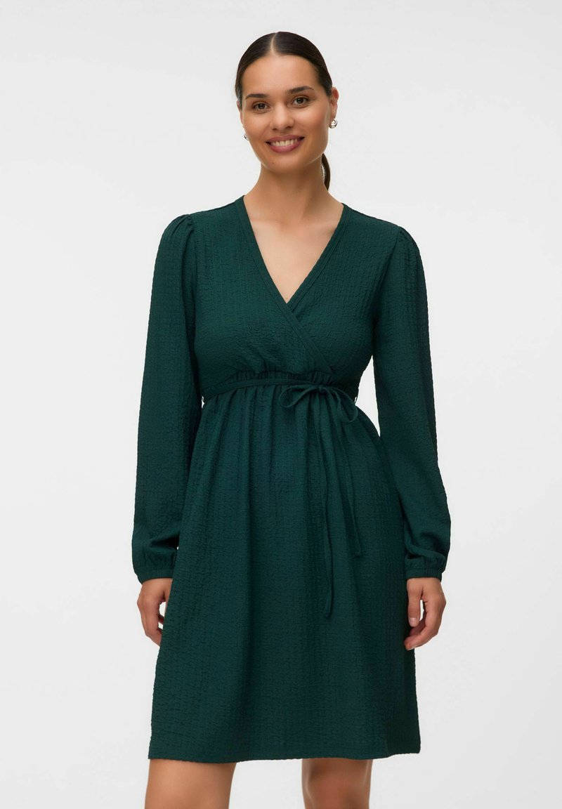 Femme souriante, debout devant un fond uni, portant une robe portefeuille à manches longues vert foncé avec un lien à la taille.