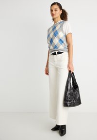 Νεαρή γυναίκα που φοράει λευκό παντελόνι, μαύρες μυτερές μπότες, άσπρο κοντομάνικο πουκάμισο και μπλε-γκρι γιλέκο αργκλάι, κρατώντας μια μαύρη υφασμάτινη τσάντα.