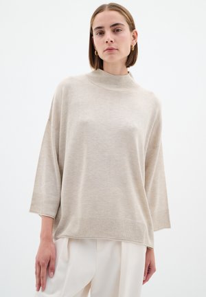 Beige Strickpullover mit hohem Kragen, weiten Ärmeln und gerippten Akzenten an Saum und Bündchen, präsentiert über hellen Hosen.