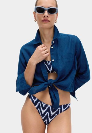 Kobieta w niebieskim bikini w wzory, zawiązana ciemnoniebieska koszula, niebieskie okulary przeciwsłoneczne i złote kolczyki koła, pozująca na jednolitym tle.