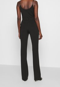 Person sedd bakifrån i en svart åtsittande ärmlös topp och högmidjade svarta byxor med lätt utsvängning och klackskor på vit bakgrund.