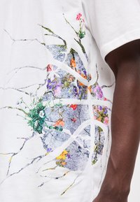 Λευκό κοντομάνικο πουκάμισο με πολύχρωμο λουλουδάτο και ραγισμένη υφή στη μία πλευρά, φορεμένο από άτομο με σκουρόχρωμο τόνο δέρματος.