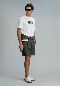 Witte katoenen t-shirt met een natuurafbeelding, olijfgroene shorts, een grijze trui rond de taille geknoopt, en witte sneakers met bruine zolen.