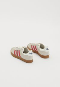 Adidași cu partea superioară din suede bej, dungi roșii, detalii albe și talpă din cauciuc maro. Include logo pe toc și un design clasic cu șireturi.