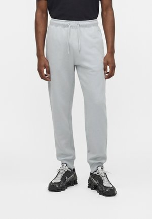 Hombre con pantalones joggers gris claro con cintura ajustable con cordón y zapatillas Nike negras y blancas, de pie sobre un fondo blanco liso.