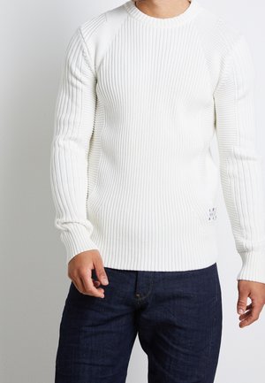 Mann trägt einen weißen, gerippten Pullover mit langen Ärmeln und dunkelblaue Jeans, steht vor einem schlichten hellen Hintergrund.