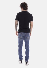 Homme avec des tatouages portant un t-shirt noir, un jean bleu et des baskets blanches, debout de dos sur un fond blanc uni.