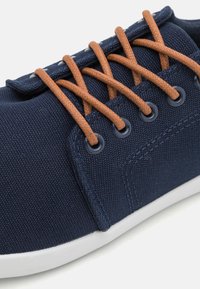 Tengerészeti vászon sneaker barna fűzőkkel, fehér gumitalppal, kerek orral és fém fűzőlyukakkal. Texturált anyagfelület megerősített varrással.