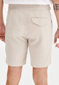 Beżowe szorty w paski wykonane z lekkiego materiału. Posiadają zapiętą kieszeń z tyłu oraz pasek w talii z pętlą. Schludny, dopasowany krój.