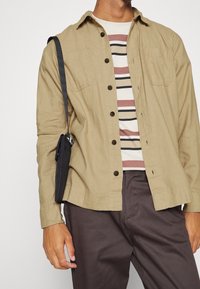 Camicia khaki con bottoni e due tasche sul petto, indossata sopra una maglietta a righe. Borsa nera con tracolla. Pantaloni scuri.