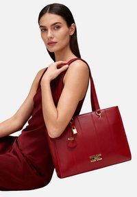 Borsa a mano in pelle rossa con cuciture verticali, dettagli in oro e chiusura con lucchetto. Presenta una forma rettangolare spaziosa e manici robusti.