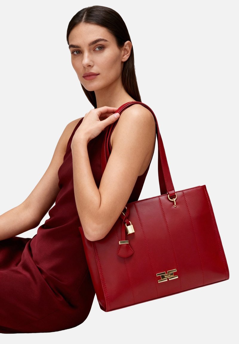 Borsa a mano in pelle rossa con cuciture verticali, dettagli in oro e chiusura con lucchetto. Presenta una forma rettangolare spaziosa e manici robusti.