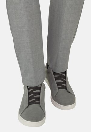 Grijze suède sneakers met zwarte veters gecombineerd met lichtgrijze formele broeken, gezien van voren op een witte achtergrond.