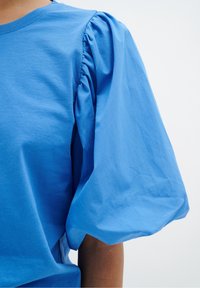 Blå bomullst-shirt med puffärmar och plisserade detaljer. Slät yta och vanlig passform, visad mot en ljus bakgrund.