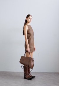 Žena u smešnoj kožnoj haljini bez rukava smeđe boje, drži odgovarajuću smeđu kožnu torbu, nosi smeđe čizme do kolena, stojeći pored jednostavnog zida.