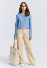 Pulover bleu deschis cu dungi, pantaloni crem cu croială largă, adidași albi, ținând o geantă tote bej cu logo negru. Texturi netede și croială lejeră.