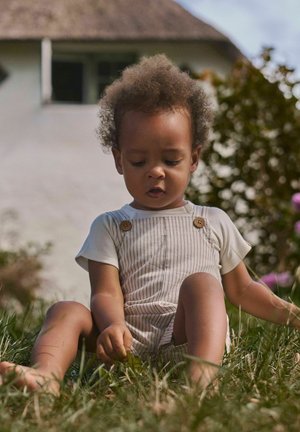 Lille barn med krøllet hår iført stribet overalls og hvid skjorte, siddende barfodet på græsset, holder et lille blad udendørs.