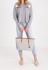 Grauer, taillierter Pullover mit rot-schwarzer Vogelstickerei, hellblaue Jeans, beigefarbene, gemusterte Tragetasche mit braunen Griffen und helle Slipper.