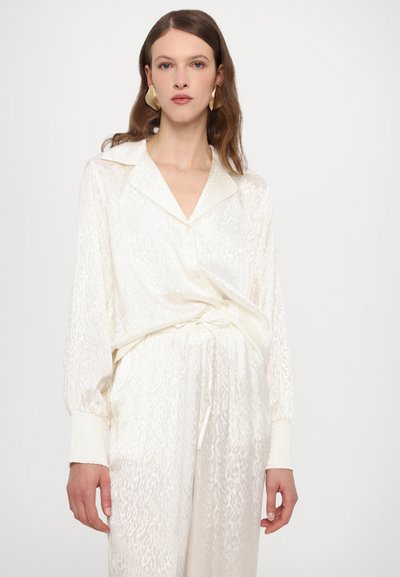 White, sheer blouse with a subtle pattern, long sleeves, and a collared neckline. Paired with matching white trousers.