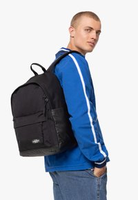 Zaino nero con corpo in tessuto e base sintetica nera lucida. Presenta numerose tasche con zip e spallacci regolabili.