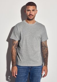 Graues Baumwoll-T-Shirt mit Rundhalsausschnitt und kurzen Ärmeln, das eine glatte Textur aufweist. Kombiniert mit blauen Jeans und dekorativen Tätowierungen an den Armen.