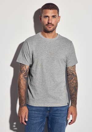Camiseta de algodón gris con cuello redondo y mangas cortas, con una textura suave. Lleva puesta con pantalones vaqueros azules y tatuajes decorativos en los brazos.