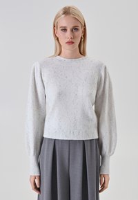 Donna bionda con orecchini ad anello dorati, che indossa un maglione a maglia leggero con macchie e maniche lunghe a pallone, abbinato a pantaloni grigi a pieghe a vita alta.