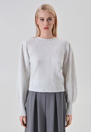 Donna bionda con orecchini ad anello dorati, che indossa un maglione a maglia leggero con macchie e maniche lunghe a pallone, abbinato a pantaloni grigi a pieghe a vita alta.
