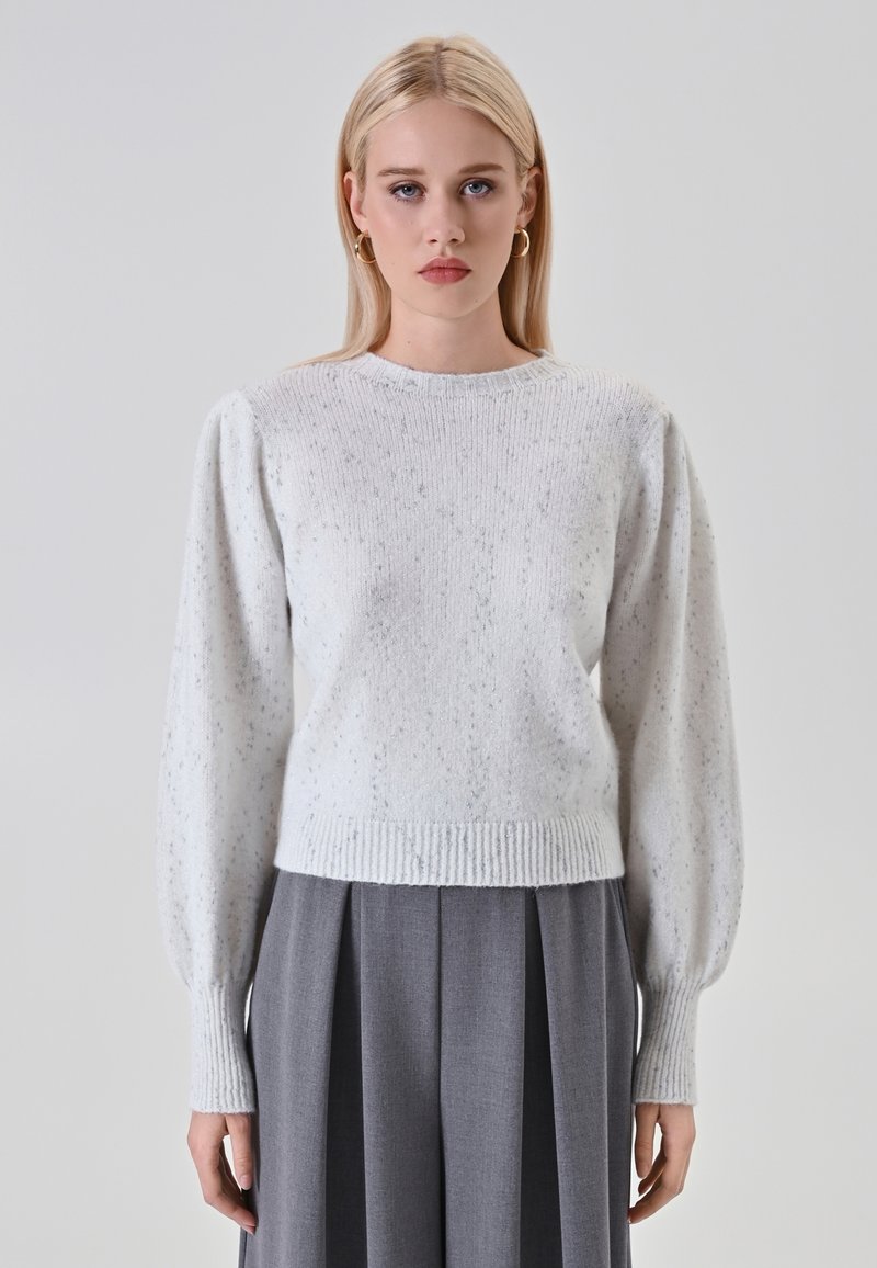 Donna bionda con orecchini ad anello dorati, che indossa un maglione a maglia leggero con macchie e maniche lunghe a pallone, abbinato a pantaloni grigi a pieghe a vita alta.