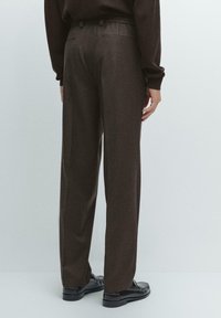 Person wearing dark brown tailored pants and black loafers, standing against a plain light background, shown from the back.