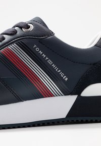 Granatowe trampki Tommy Hilfiger z czerwonymi, białymi i czarnymi paskami, czarnymi sznurówkami i białą podeszwą.