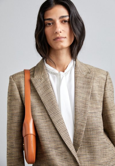 Brun ternet blazer med et struktureret stof, hvid bluse underneath og en orange lædertaske. Blazeren har en klassisk skæring og slåede kraver.