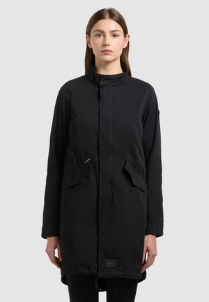 Donna con capelli castani lisci, che indossa un lungo cappotto nero con tasche frontali a pattina, in piedi davanti a uno sfondo chiaro e uniforme.