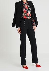 Blazer nero su misura sopra una blusa floreale con fiori rossi e rosa. Pantaloni neri aderenti con dettagli a bottoni. Tacchi alti rossi a punta.