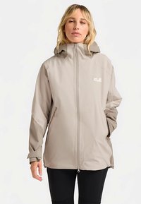 Giacca impermeabile beige chiaro con zip frontale, cappuccio e polsini regolabili. Tasche laterali con zip e marchio minimalista sul petto in alto a sinistra.