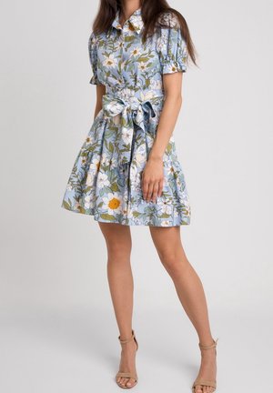 Frau trägt ein hellblaues Blumenkleid mit Puffärmeln und einem großen Schleifengürtel, kombiniert mit beigen Sandalen mit Knöchelriemen und hohen Absätzen.