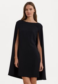 Musta mekko, jossa on tiukka siluetti ja viittamainen päällinen. Su smooth-kosketus, venepääntie ja lyhyt pituus.