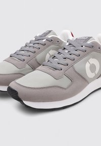Baskets de sport grises avec un mélange de matériaux lisses et texturés, arborant un logo blanc, des lacets gris et une semelle en caoutchouc noire.