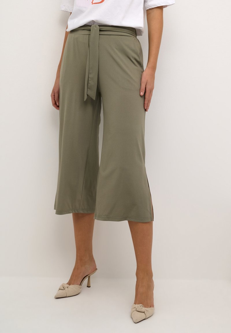 Vrouw in olijfgroene, cropped wijde broek met een strikceintuur en beige, puntige hakschoenen op een effen achtergrond.