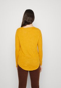 Långärmad topp i melerad senapsgul färg, med en rundad halsringning och en böjd fåll. Matchad med bruna ribbade byxor.