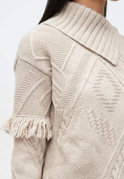 Beige neulepaita, jossa on teksturoidut geometriset kuviot, hapsutetut lyhyet hihat ja leveä resorikaulus henkilöllä, jolla on tummat hiukset.