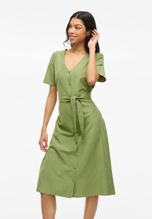 Femme souriante, portant une robe midi olive à manches courtes et ceinture, posant contre un fond blanc uni.